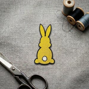 May include: A yellow embroidered patch in the shape of a rabbit silhouette, featuring long ears and a small white circular tail. The patch is positioned on a grey fabric surface alongside metal scissors and three spools of sewing thread.