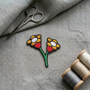 Peut inclure: Un écusson brodé représentant deux fleurs stylisées avec des pétales jaunes, des centres blancs et des bases rouges, reliées par une tige verte unique. L'écusson est posé sur un tissu gris près de ciseaux en métal et trois bobines de fil.