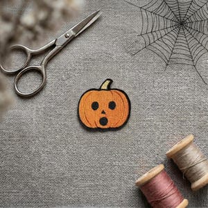 May include: An embroidered pumpkin patch featuring an orange jack-o'-lantern with a surprised expression, black circular eyes, and a black mouth. The patch is positioned near silver metal scissors and two wooden spools of thread on a grey fabric.