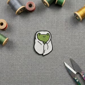 Peut inclure: Un écusson brodé représentant une grenouille verte enveloppée dans une couverture blanche avec des contours noirs. L'écusson est entouré de plusieurs bobines de fil en bois gris, vert et jaune, ainsi qu'une paire de ciseaux en métal sur un tissu gris.