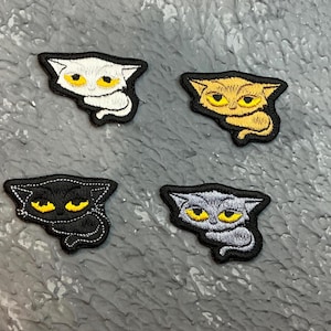 May include: Four embroidered cat patches in white, gold, black, and grey. Each patch features a cat face with yellow eyes and a black outline. The patches are on a textured grey surface.