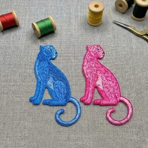 May include: Two embroidered patches of sitting panthers, one blue and one pink, with curled tails. The patches are on a gray fabric surface, with spools of thread and gold scissors in the background. The patches are detailed with intricate stitching.