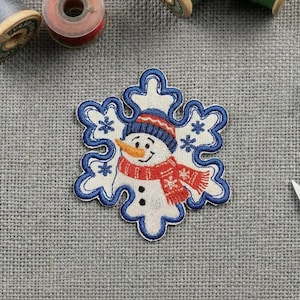 Peut inclure: Un écusson brodé en forme de flocon de neige avec une bordure bleue. Le centre représente un bonhomme de neige portant un bonnet en tricot bleu et une écharpe rouge à motifs de flocons blancs. Il a un nez en carotte orange et deux boutons noirs.