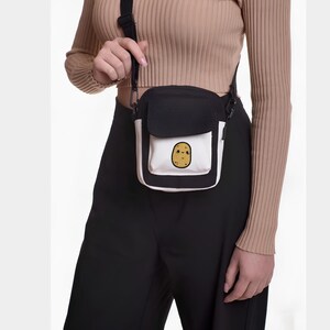 Op de afbeelding: Zwarte en witte crossbody tas met een klep. De tas heeft een witte zak met een cartoon aardappel illustratie. Het wordt over de schouder gedragen met een zwarte band.