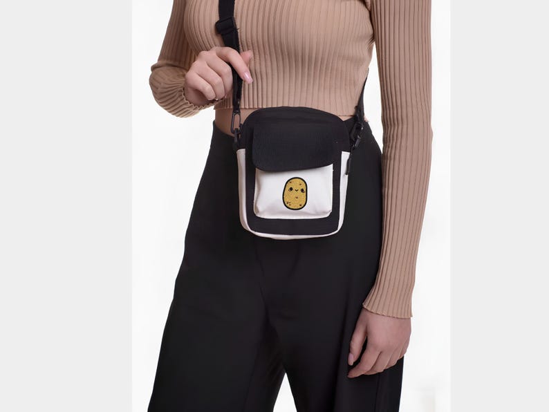 Op de afbeelding: Zwarte en witte crossbody tas met een klep. De tas heeft een witte zak met een cartoon aardappel illustratie. Het wordt over de schouder gedragen met een zwarte band.