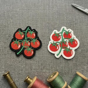 May include: Two embroidered tomato patches, each depicting a cluster of red tomatoes with green stems. One has a black border, the other a white border. Scissors and spools of thread are also visible.