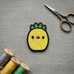 Peut inclure: Un écusson ananas jaune avec un contour noir et un dessus vert. Le patch a un motif de grille cousu et trois petits points noirs pour les yeux. Ciseaux dorés et bobines de fil en arrière-plan.
