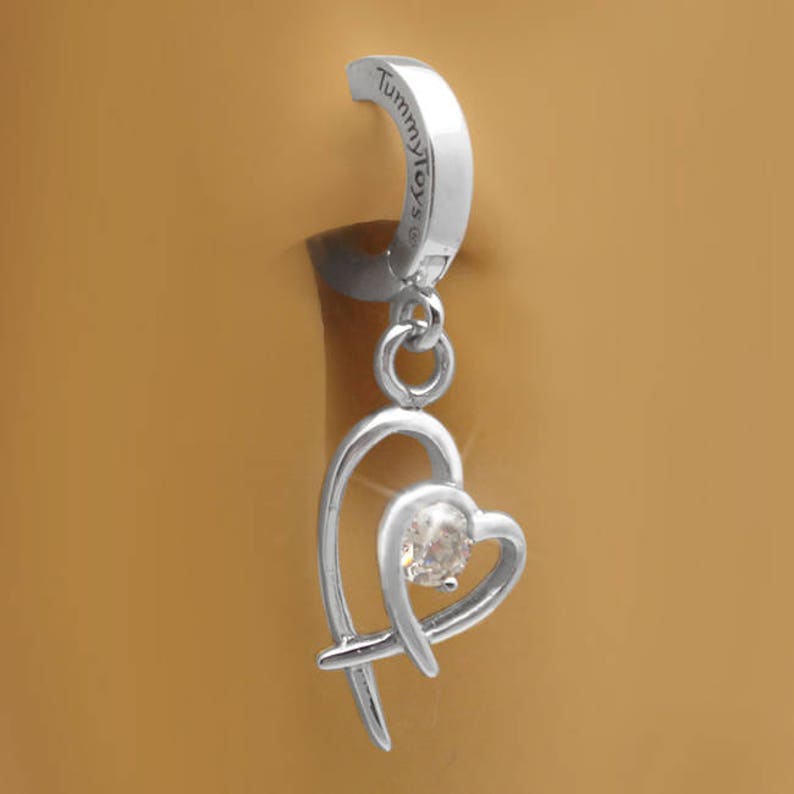Puede incluir: Anillo de ombligo plateado con un colgante de coraz&oacute;n que cuelga con un cristal transparente. El anillo est&aacute; grabado con las palabras "Tummy Toys".