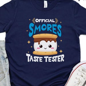 May include: A navy blue t-shirt with a white graphic of a smiling s'more with the text "OFFICIAL SMORES TASTE TESTER".