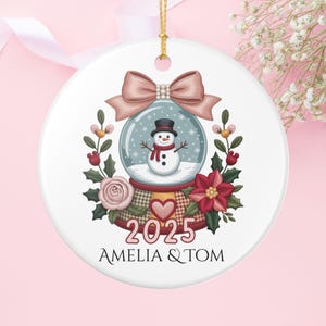 Könnte beinhalten: Runder, weißer Weihnachtsschmuck mit einem Schneemann in einer Schneekugel. Der Schmuck hat eine rosa Schleife, florale Akzente und den Text "2025 AMELIA & TOM". Ein goldener Faden ist zum Aufhängen befestigt.