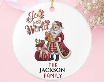May include: A white ceramic Christmas ornament with a gold hanging loop. The ornament features a Santa Claus illustration reading a book, a red gift bag, and the text "Joy to the World" and "The Jackson Family."