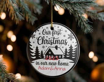 May include: Round Christmas ornament with the text "Our first Christmas" and "in our new home 2023". The ornament features a house silhouette, snowflakes, and pine trees. The names "Adam & Anna" are printed at the bottom.