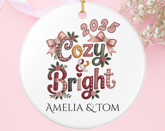 May include: A round, white ceramic Christmas ornament with the words "Cozy & Bright" and the year "2025" in a decorative font. The ornament includes the names "AMELIA & TOM" at the bottom. The design features bows, floral accents, and a gold hanging loop.