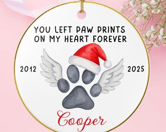 May include: A white ceramic Christmas ornament with a gold rim and a gold string for hanging. The ornament features a paw print with angel wings, a Santa hat, and the text "YOU LEFT PAW PRINTS ON MY HEART FOREVER". The name "Cooper" is written in red.