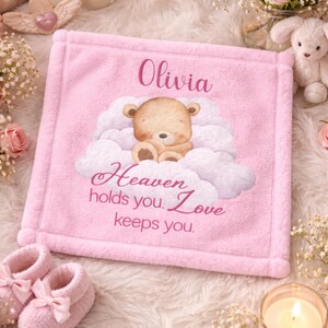Könnte beinhalten: Eine rosa Babydecke mit dem Namen "Olivia" oben aufgedruckt. Die Decke zeigt eine Teddybär-Illustration auf einer Wolke mit dem Text "Heaven holds you. Love keeps you."