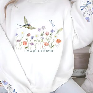 May include: White sweatshirt with a floral design and a bluebird flying over a field of flowers. The text "I'm a wild flower" is printed below the flowers.