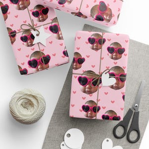 Könnte beinhalten: Rosa Geschenkpapier mit einem sich wiederholenden Muster eines Babys mit herzförmiger Sonnenbrille. Die Geschenke sind mit Bindfaden gebunden und mit weißen Papierauschnitten verziert. Ein Knäuel Bindfaden und eine Schere sind ebenfalls zu sehen.