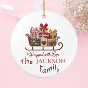 Könnte beinhalten: Weißes Keramik-Weihnachtsornament mit goldenem Band. Das Ornament zeigt einen Schlitten voller Geschenkpakete in Rosa, Rot und Grün. Der Text lautet "Wrapped with Love" und "The JACKSON Family".