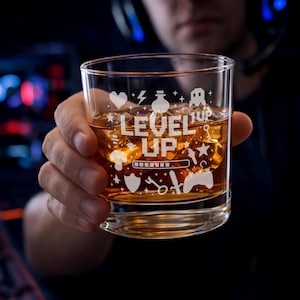 Könnte beinhalten: Ein klares Glas mit bernsteinfarbener Flüssigkeit und Eiswürfeln. Das Glas ist mit den Worten "LEVEL UP" und verschiedenen Gaming-Symbolen graviert. Das Glas wird von einer Person gehalten.