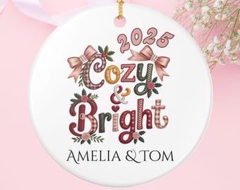 May include: A white ceramic Christmas ornament with the words "Cozy & Bright" and the year 2025 in a decorative font. The ornament has a gold string for hanging and the names "AMELIA & TOM" are printed at the bottom.