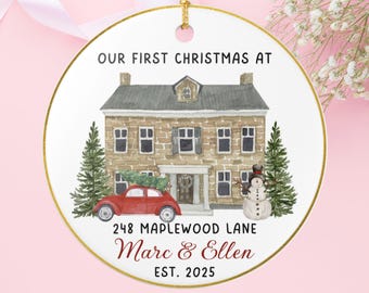 May include: A circular Christmas ornament with a gold edge. It displays a watercolour illustration of a house, a red car with a Christmas tree, and a snowman. Text reads "OUR FIRST CHRISTMAS AT", address, names, and the year 2025.