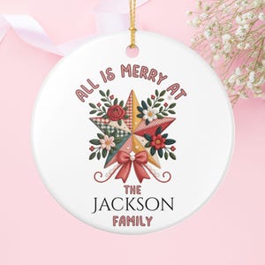 Könnte beinhalten: Weißer Keramik-Weihnachtsschmuck mit goldenem Band. Das Ornament zeigt ein Stern-Design mit floralen Akzenten und dem Text "ALL IS MERRY AT THE JACKSON FAMILY". Der Stern besteht aus verschiedenen gemusterten Stoffen.