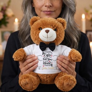 Könnte beinhalten: Ein brauner Teddybär mit schwarzer Fliege und weißem T-Shirt. Das T-Shirt hat aufgedruckten Text und Daten. Der Bär wird von einer Person mit grauem Haar gehalten.