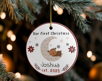 Può includere: Ornamento natalizio rotondo con sfondo bianco e bordo marrone. L'ornamento presenta un orsetto che dorme su una luna crescente, fiocchi di neve e il testo "Our first Christmas" e "Joshua est. 2025".