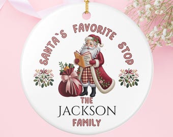 May include: A white ceramic Christmas ornament with a gold string. The ornament features Santa Claus reading a book, a gift bag, and the text "Santa's Favorite Stop" and "The Jackson Family" in a festive font.