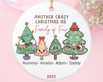 May include: White ceramic Christmas ornament with a gold string. The ornament features four cartoon Christmas trees with faces, the text "ANOTHER CRAZY CHRISTMAS AS Family of Four", and the names "Mommy- Amelia- Adam- Daddy". The year "2025" is also printed.