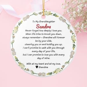 Könnte beinhalten: Runder weißer Keramikschmuck mit goldener Aufhängeschnur. Er hat einen floralen Rand und eine Botschaft an "Sandra" von "Oma". Der Text ist in Schwarz und Rot gehalten und drückt Liebe und Unterstützung aus.