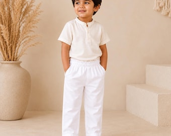 May include: A short-sleeved cream-colored shirt with a partial button-down placket and a mandarin collar, paired with plain white trousers featuring an elasticized waistband. The outfit is clean, simple, and constructed from lightweight fabric.