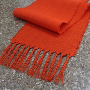 May include: Orange table runner with a fringe edge. The runner is made of a woven fabric and has a subtle texture.