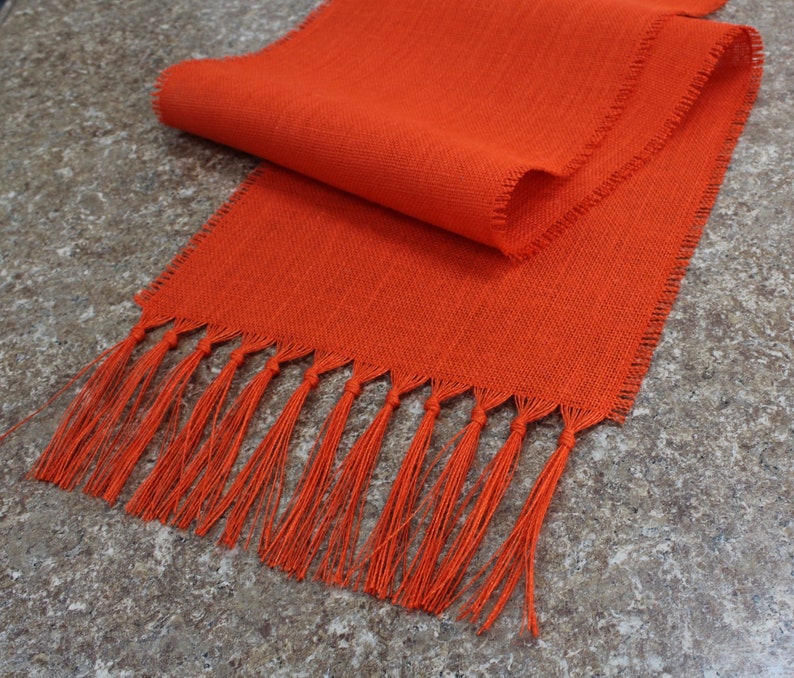 May include: Orange table runner with a fringe edge. The runner is made of a woven fabric and has a subtle texture.