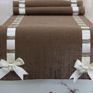 May include: A brown burlap table runner with white satin ribbon trim and two white satin bows at the ends.