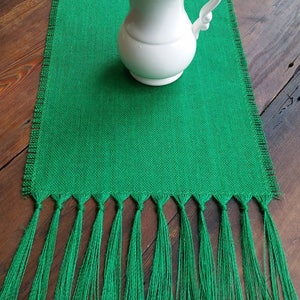 May include: A green burlap table runner with fringe. The runner is 14 inches wide and has a white pitcher on top of it.