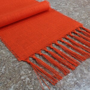 May include: Orange linen table runner with fringe. The runner is rolled up on the left side of the image.