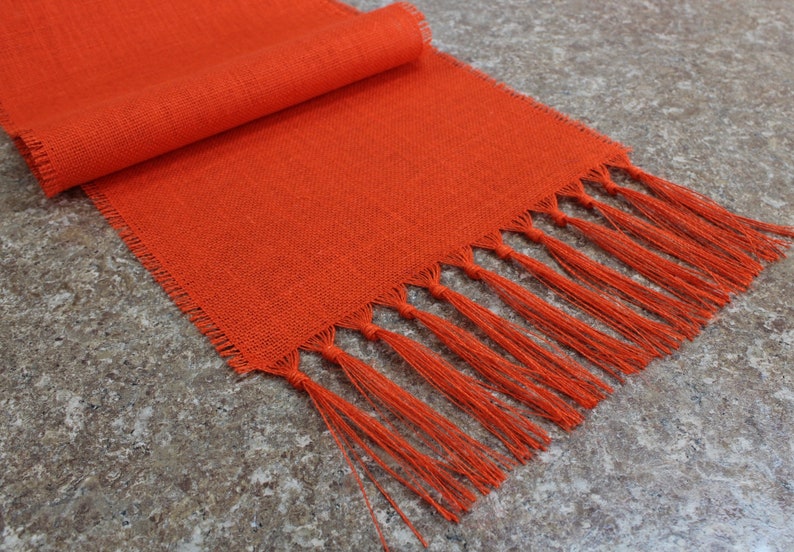 May include: Orange linen table runner with fringe. The runner is rolled up on the left side of the image.