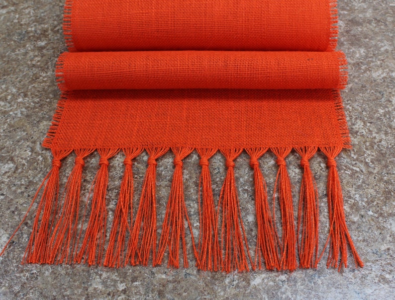 May include: Orange burlap table runner with fringe. The runner is folded in half and has a woven texture.