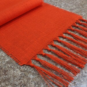 May include: Orange linen table runner with fringe. The runner is woven with a subtle texture and has a tassel fringe on both ends.