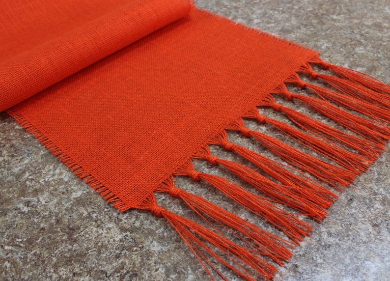 May include: Orange linen table runner with fringe. The runner is woven with a subtle texture and has a tassel fringe on both ends.
