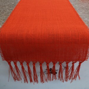 May include: Orange burlap table runner with fringe. The runner is made of a woven fabric with a rough texture. The fringe is made of the same fabric and is about 1 inch long.