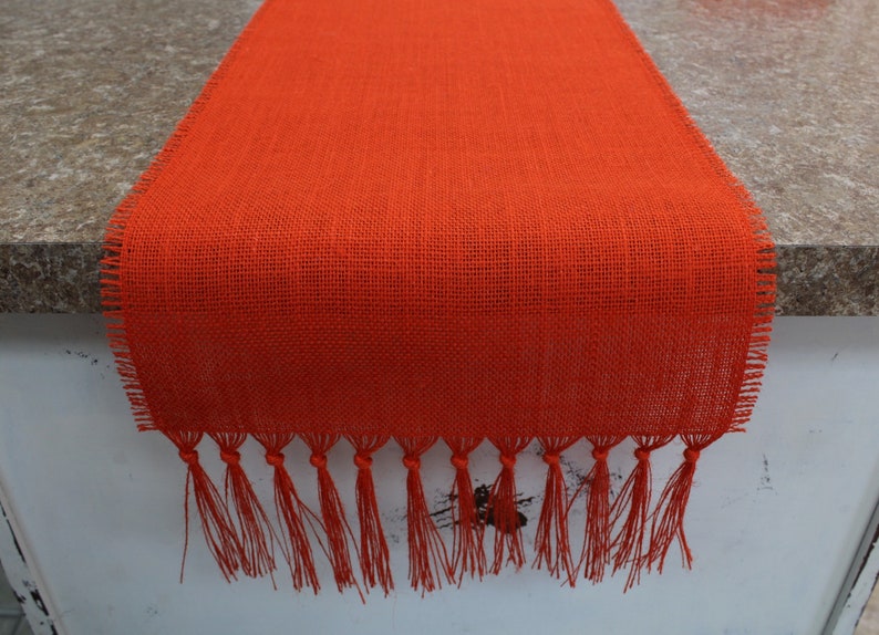 May include: Orange burlap table runner with fringe. The runner is made of a woven fabric with a rough texture. The fringe is made of the same fabric and is about 1 inch long.