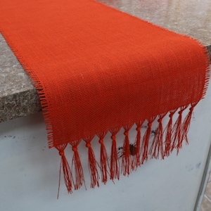 May include: Orange burlap table runner with fringe. The runner is made of a woven fabric with a rough texture. The fringe is made of the same fabric and is tied in knots.