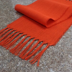 May include: A bright orange table runner with a fringe border. The runner is made of a woven fabric and has a subtle texture.