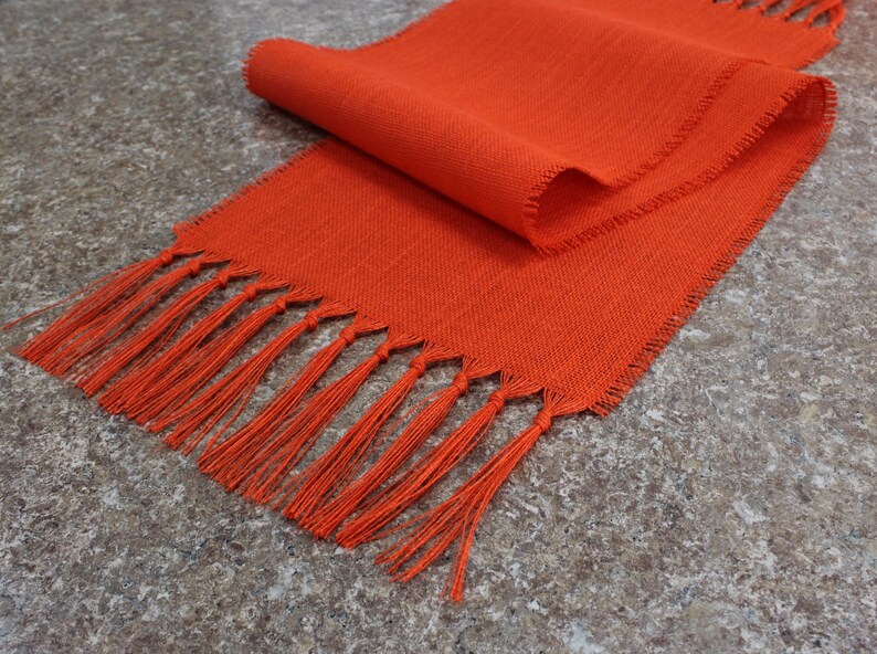 May include: A bright orange table runner with a fringe border. The runner is made of a woven fabric and has a subtle texture.