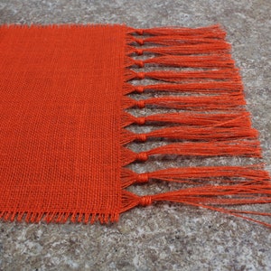 May include: Orange burlap table runner with a fringe edge. The runner is made of a woven fabric with a rough texture.