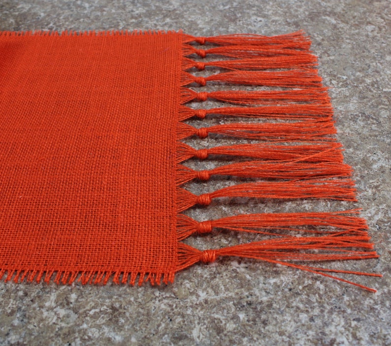 May include: Orange burlap table runner with a fringe edge. The runner is made of a woven fabric with a rough texture.