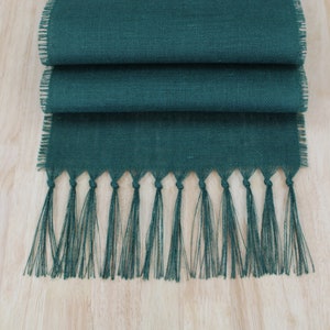 May include: A dark green burlap table runner with fringed edges. The runner is folded in thirds and has a tassel fringe.