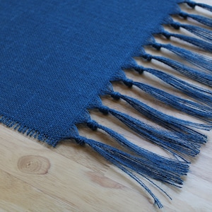 May include: A blue woven table runner with fringe on the ends. The runner is on a light wood surface.